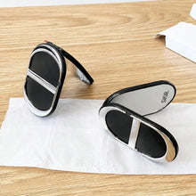 Two oval-shaped black and silver compact mirrors with a glossy finish, displayed on a white tissue paper over a light wood grain surface.