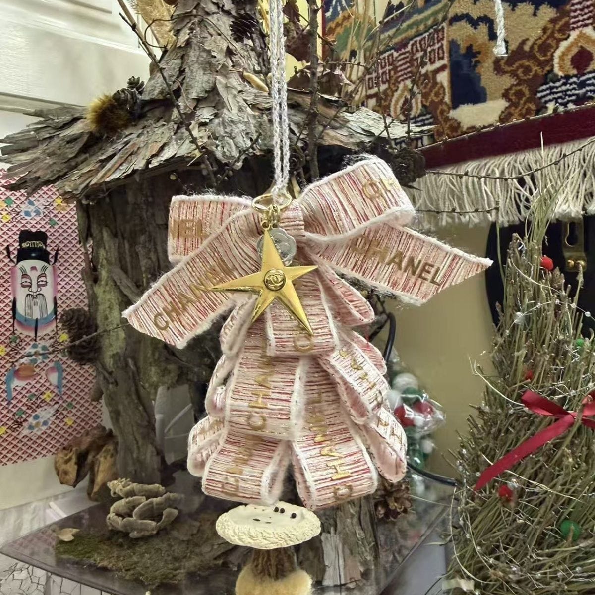 A decorative hanging ornament shaped like a stylized angel, crafted from ribbon-like strips printed with the Chanel wordmark and a gold star at the center, shown hanging in a styled indoor display.
